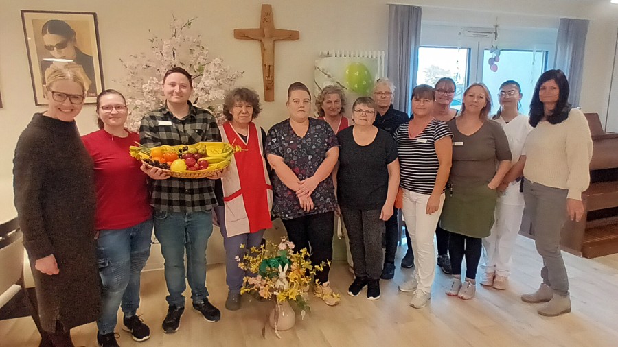 Eine Gruppe von zwölf Personen posiert lächelnd, eine Person hält einen Korb mit frischem Obst in der Hand.